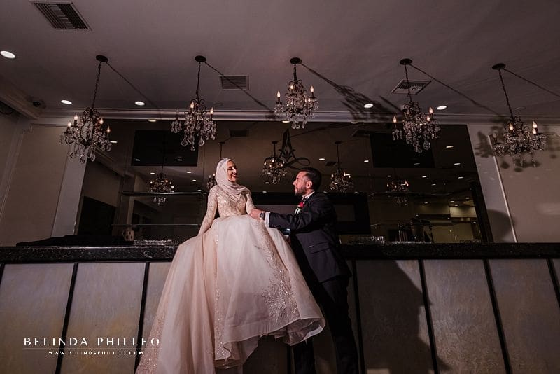 Bride sits atop bar and admires her groom during Brandview Ballroom wedding.