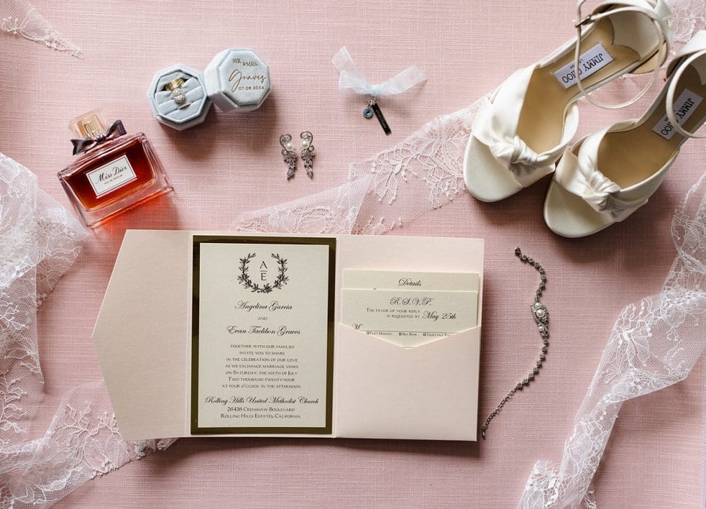 A flatlay of bridal deatils with blush and ivory tones, including a blush and gold invitation suite, ivory Jimmy Choo shoes, and Chloe perfume