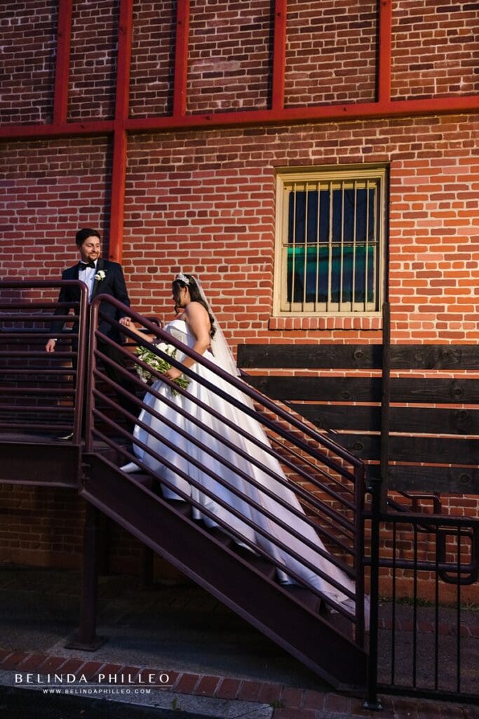 Groom leads his bride by the hand up the stairwell in front of a brick wall