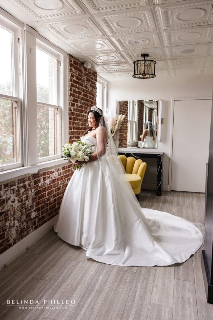 Portrait of a bride standing near a window, admiring her bouquet in the bridal suite at The Charleston in Fullerton, CA